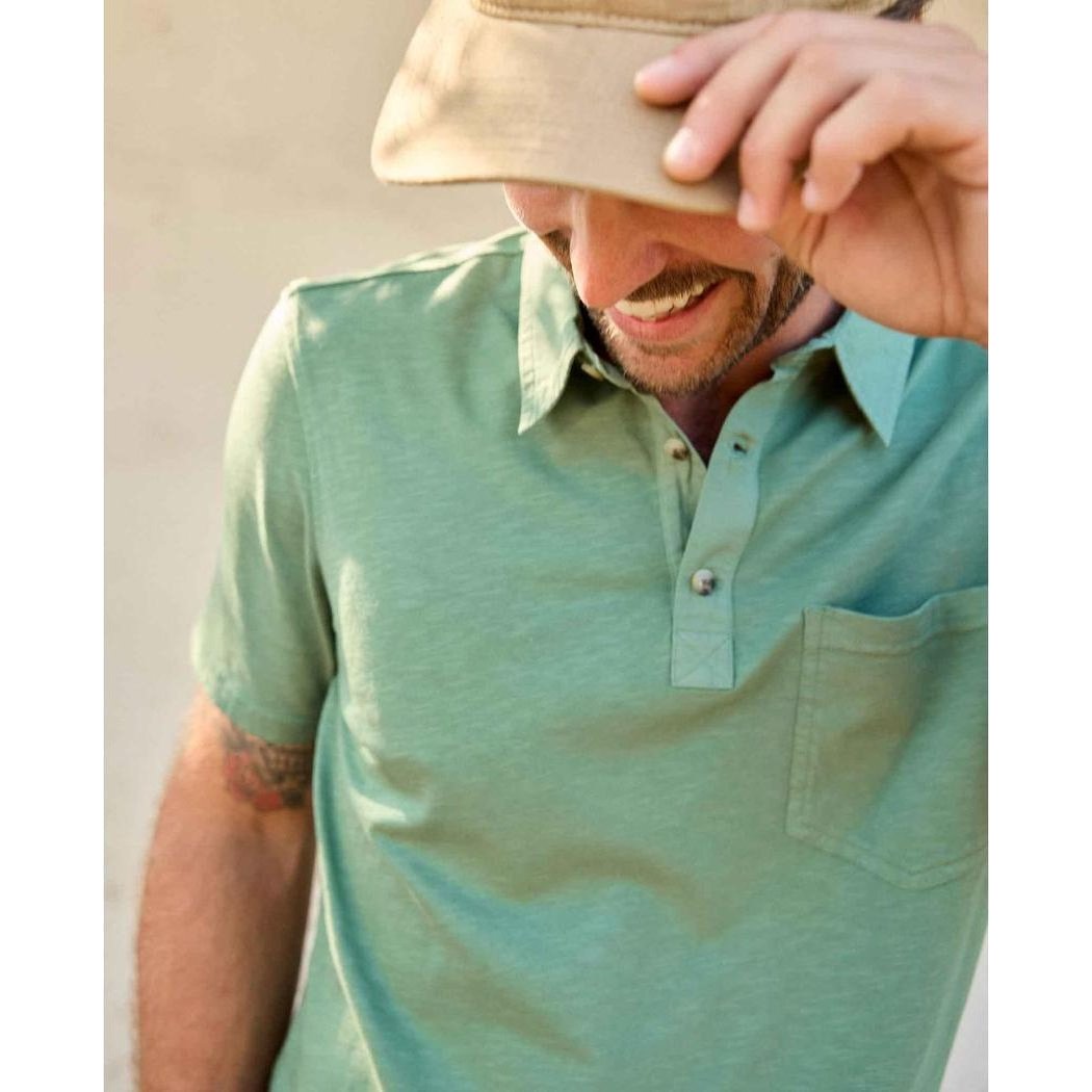 A man wearing the Toad & Co Men's Primo Short Sleeve Polo in Ivy adjusts his hat, showing the front pocket and button placket.