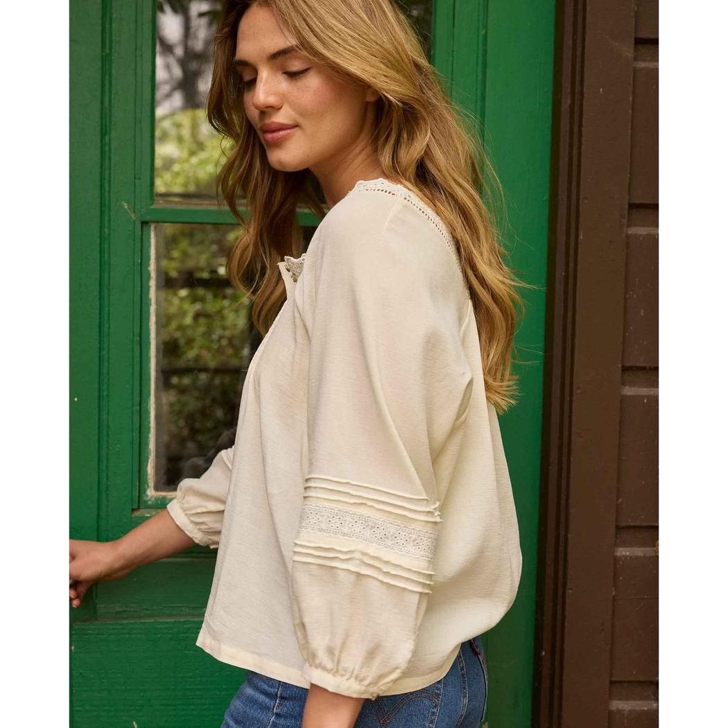 A woman wears the Toad & Co Women's Manzana Peasant LS Shirt in Egret, showing the lace detail on the sleeve and the green door background.