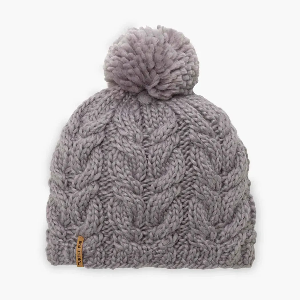A front view of the Turtle Fur Women's Merino Wool Millie hat in Black with a pom pom and a small tag on the side against a white background.