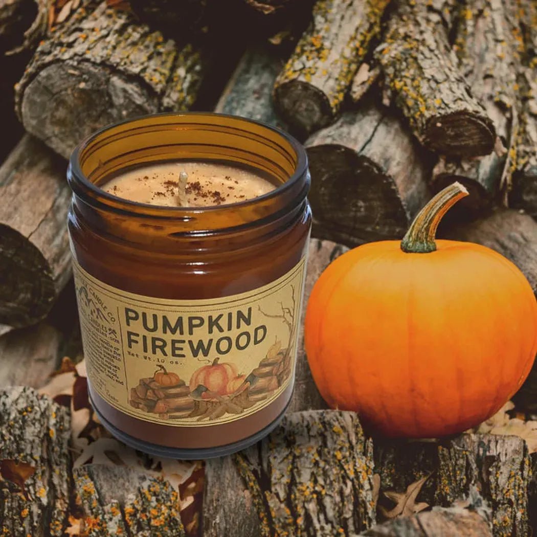 A 4 oz Pumpkin Firewood candle sits next to a small orange pumpkin on a bed of firewood logs.