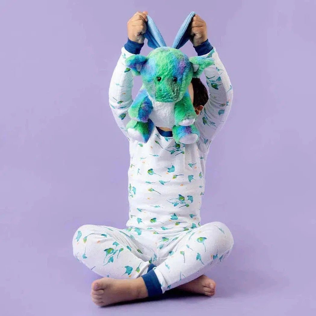 A child sits cross-legged on a purple background wearing white pajamas with green leaves, holding a blue and green Warmies Baby Dragon plush above their head.