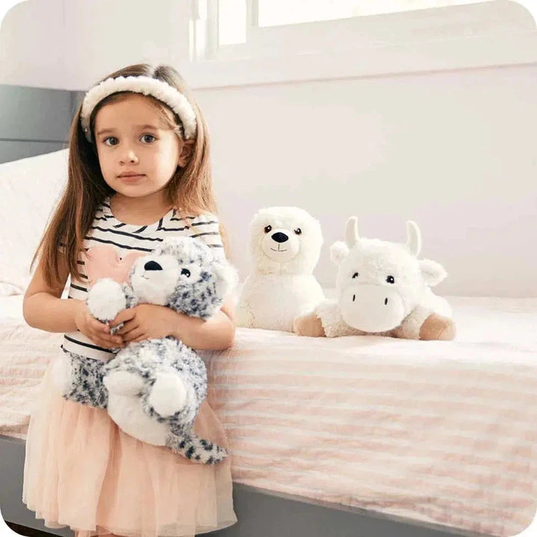 A little girl in a striped top and tulle skirt holds a Warmies Buffalo plush while two more sit on a bed with a light pink striped bedspread.