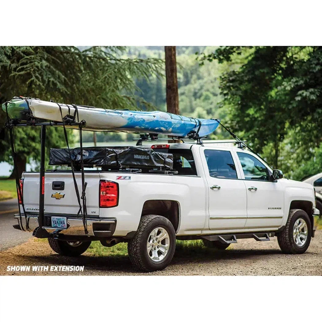 A white Chevrolet truck with a Yakima LongArm rack system carrying a blue and white kayak, parked on a gravel path with trees in the background.