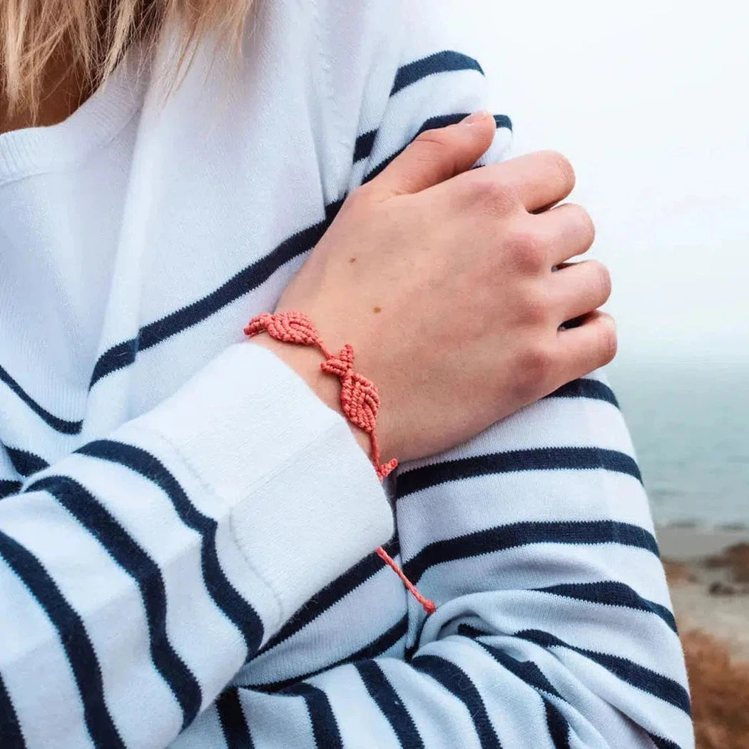 A hand wearing a Coral 4Ocean 4Fish bracelet on a striped shirt with a beach background.