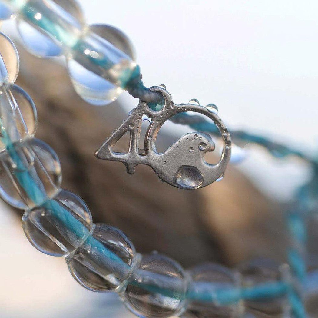A close-up of the 4Ocean Dolphin bracelet with clear beads and an aqua cord, showing the dolphin charm against a blurred outdoor background.