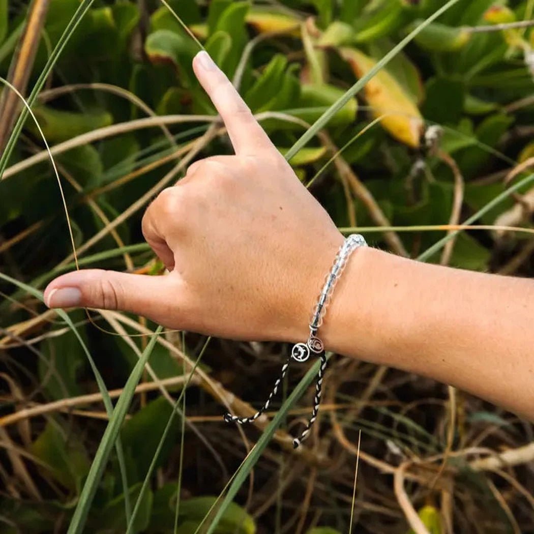 A hand wearing the 4Ocean Orca Beaded Bracelet makes a shaka sign against green foliage.
