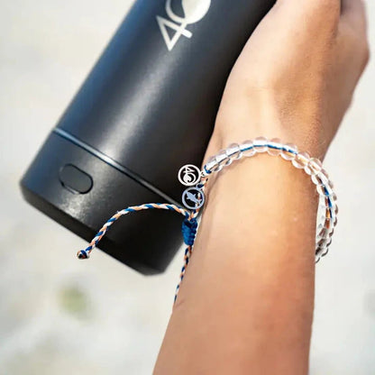 A hand holds a black 4Ocean bottle while wearing the 4Ocean Reef Shark Beaded Bracelet, showing its blue and white design.