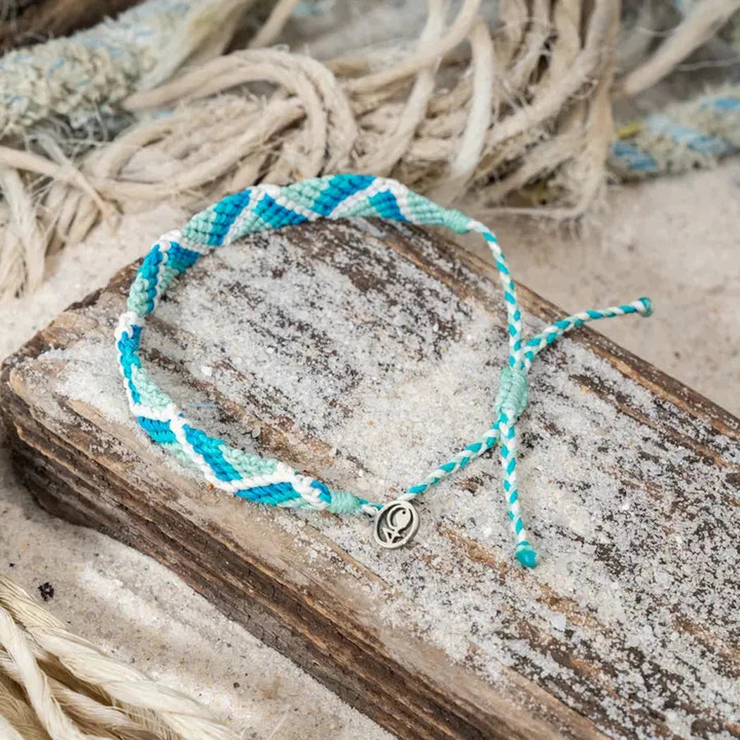 A mint glow 4Ocean Riptide Bracelet rests on a sandy wooden plank with rope in the background.