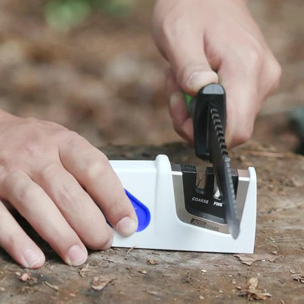 A person uses the AccuSharp Compact Pull-Through Sharpener on a tree stump in a wooded area.