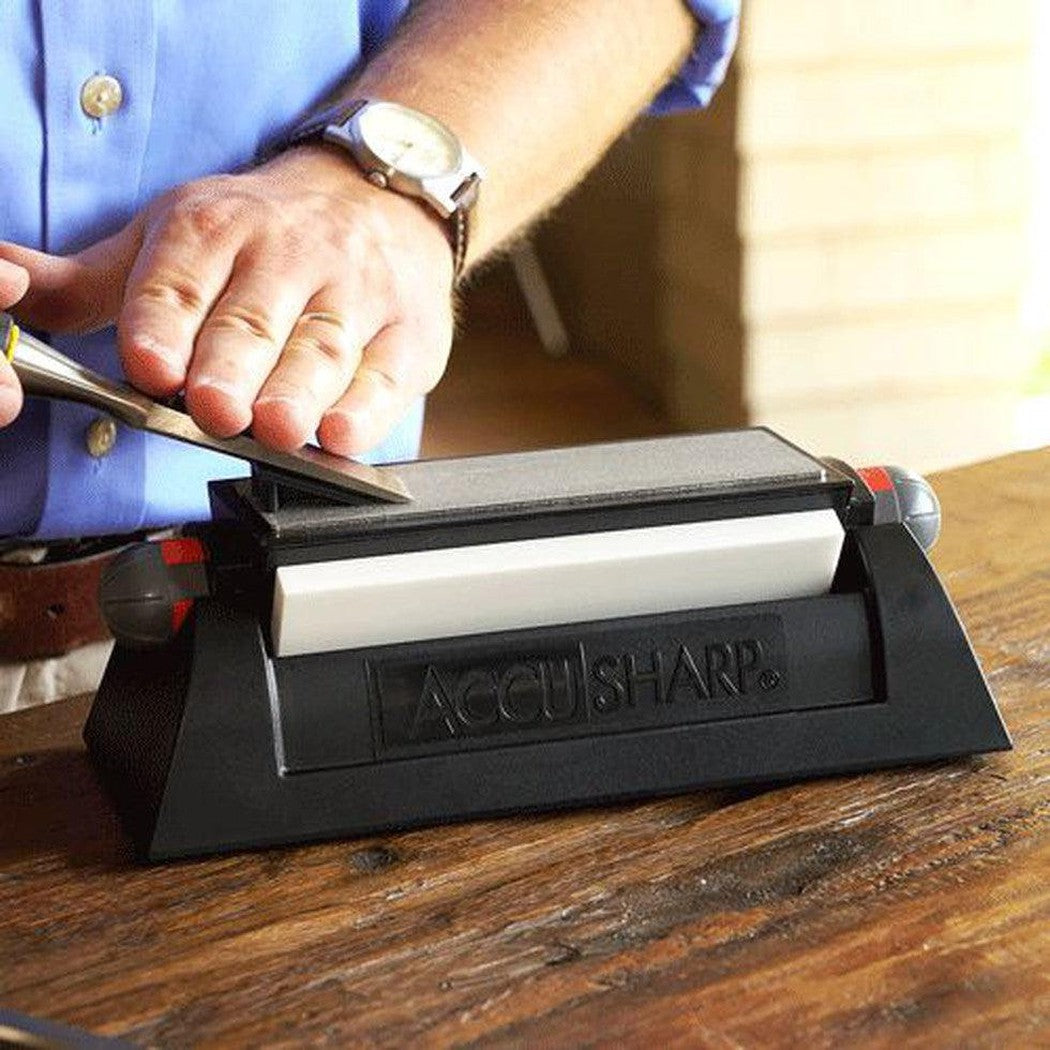 A person in a blue shirt uses the AccuSharp Tri-Stone Sharpening System on a wooden table with a brick wall background.