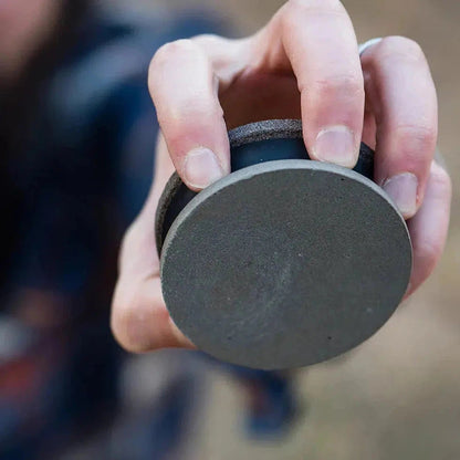 A hand holds the Adler DUAL Axe Sharpener 80/220, showing its charcoal/graphite top and black base from a close-up angle.