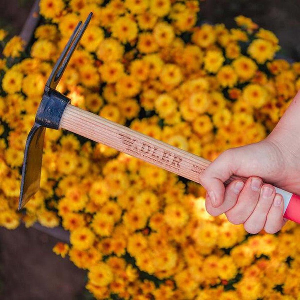 A hand holds the Adler Lily Garden Hoe with a green handle against a background of yellow flowers.