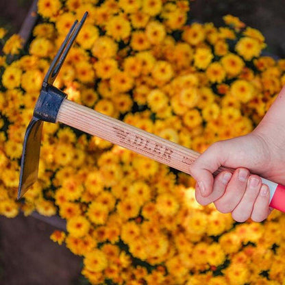 A hand holds the Adler Lily Garden Hoe with a green handle against a background of yellow flowers.