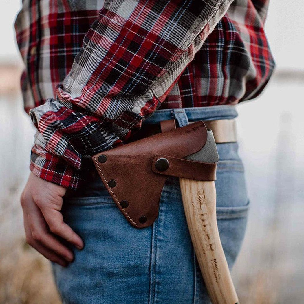 A person in a plaid shirt and blue jeans wears the Adler Scout Hatchet in a brown leather sheath at their hip, with a blurred outdoor background.