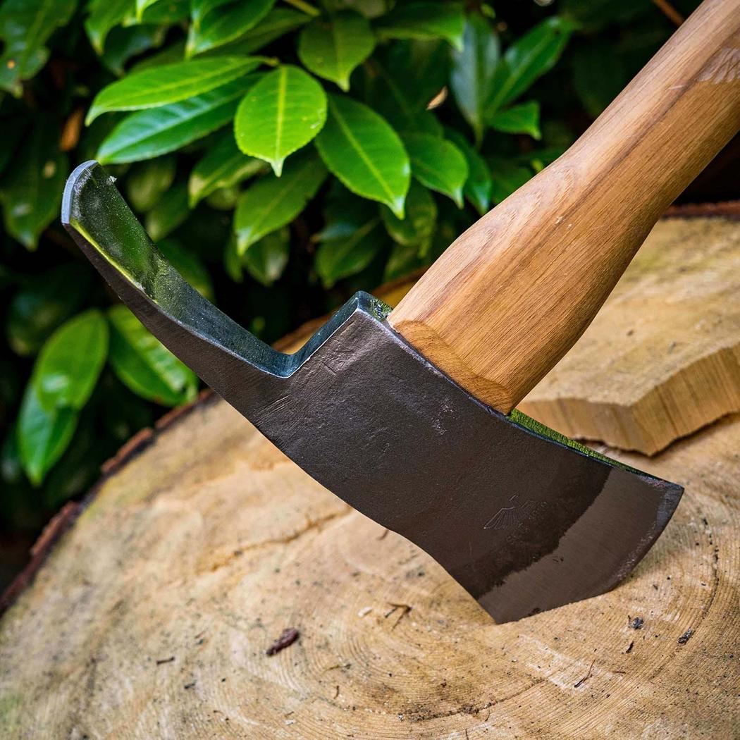 A close-up of the Adler The Pulaski Axe with its wooden handle and charcoal-colored blade resting on a tree stump against green foliage.