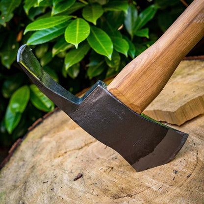 A close-up of the Adler The Pulaski Axe with its wooden handle and charcoal-colored blade resting on a tree stump against green foliage.