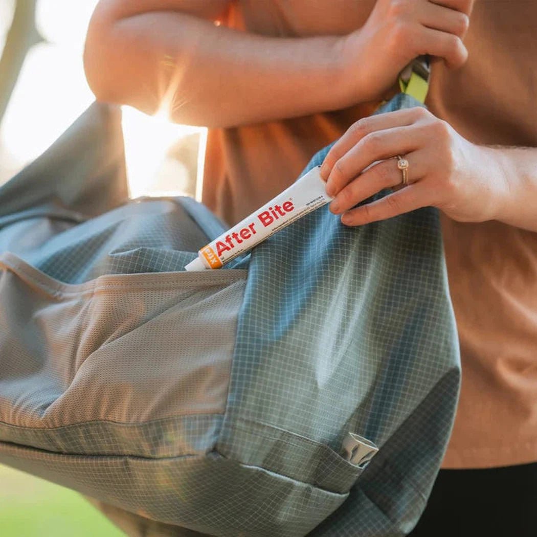A person puts After Bite Xtra in a blue backpack pocket with sunlight in the background.