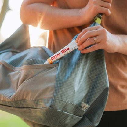 A person puts After Bite Xtra in a blue backpack pocket with sunlight in the background.