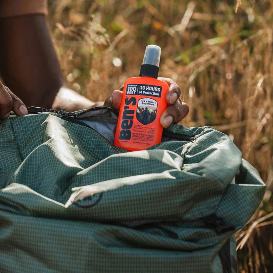 A hand holds Ben's 100 Tick and Insect Repellent Pump over a green backpack in a grassy outdoor setting.