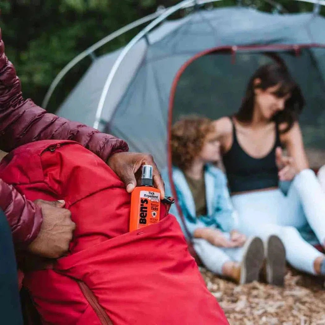 A person holds a red bag with the Adventure Medical Kits Bens Tick 3.4oz in front of a tent with two people inside.