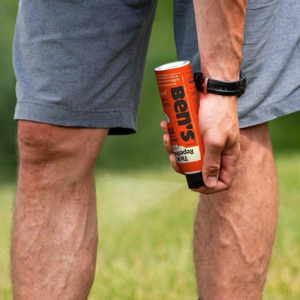 A person in gray shorts holds an orange Ben's Tick Repellent can in a grassy outdoor setting.