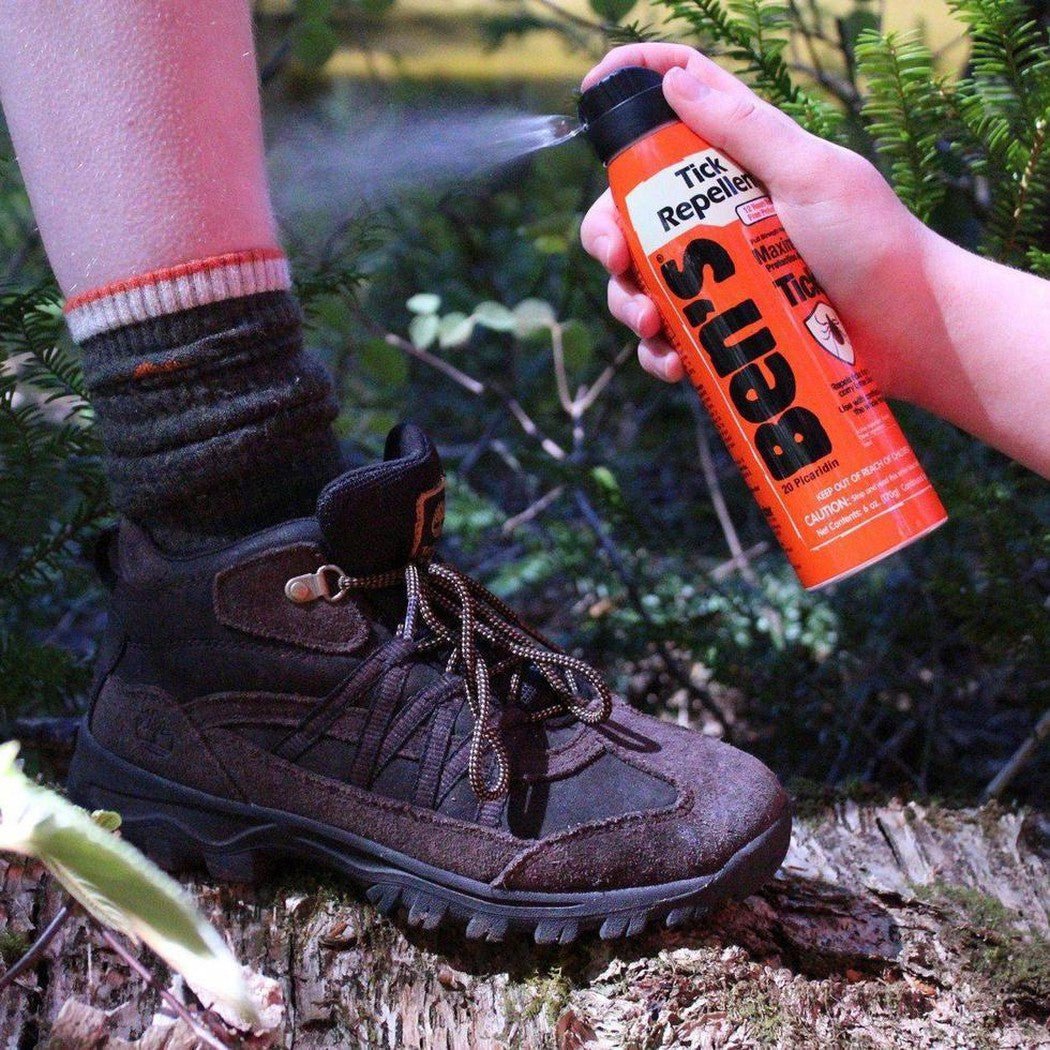 A hand sprays Ben's Tick Repellent on a hiker's leg near a brown hiking boot on a log in a forest.