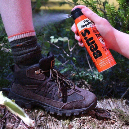 A hand sprays Ben's Tick Repellent on a hiker's leg near a brown hiking boot on a log in a forest.