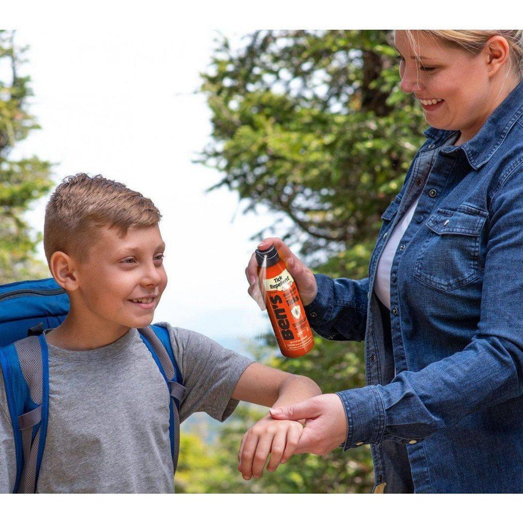 A woman sprays Bens Tick Repellent Eco-Spray on a boy's arm in a forested outdoor setting.