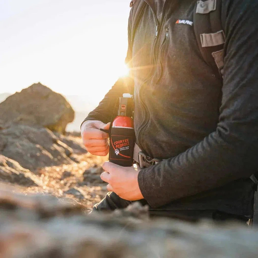 A person in a charcoal jacket holds the Adventure Medical Kits Counter Assault 32-Foot Bear Spray with Holster while sitting on rocky terrain with a sunlit background.
