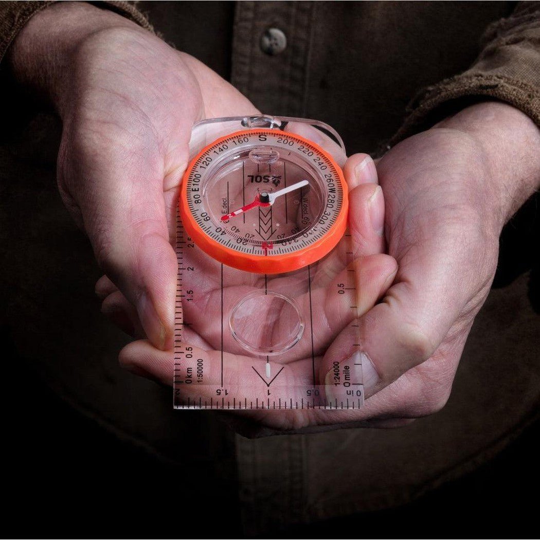 A person holds the Adventure Medical Kits Deluxe Map Compass with an orange bezel and clear base, showing the compass face and ruler features.