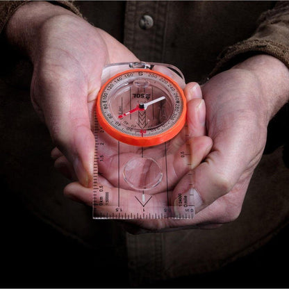 A person holds the Adventure Medical Kits Deluxe Map Compass with an orange bezel and clear base, showing the compass face and ruler features.