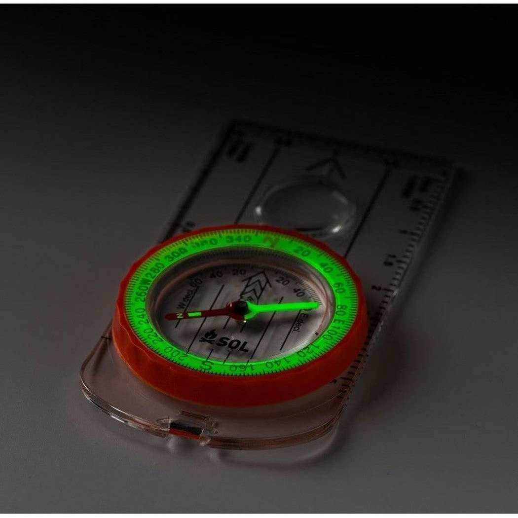 A close-up of the Adventure Medical Kits Deluxe Map Compass with its green-lit dial and orange bezel resting on a transparent base.