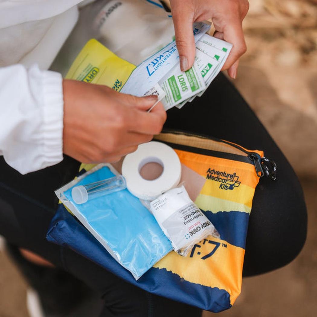 A person holds small packets over an Adventure Ready Brands AMK Ultralight/Watertight .7 first aid kit with a roll of tape and blue gauze visible.