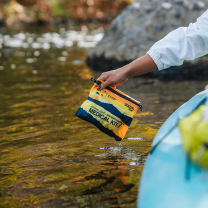 A hand holds the Adventure Ready Brands AMK Ultralight/Watertight .7 medical kit over a stream near a blue kayak.