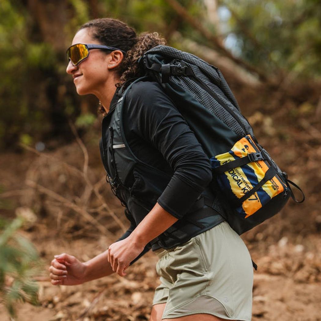 A woman hiking outdoors wears the Adventure Ready Brands AMK Ultralight/Watertight .7 first aid kit on her backpack.