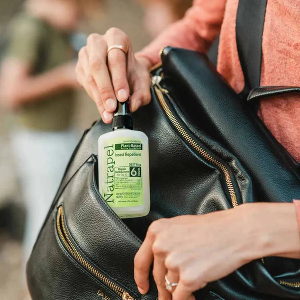 A person in a coral top holds a black backpack with a Natrapel insect repellent bottle in its pocket, showing the pump and label clearly.