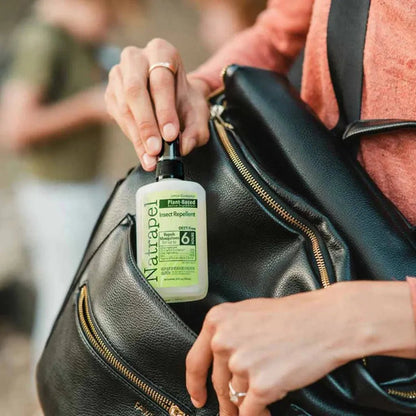 A person in a coral top holds a black backpack with a Natrapel insect repellent bottle in its pocket, showing the pump and label clearly.