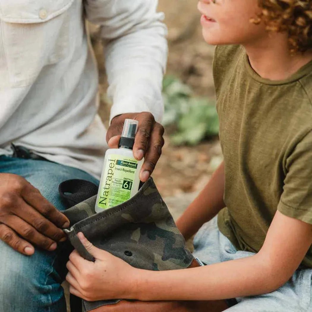 A person puts Natrapel Lemon Eucalyptus Pump into a camouflage bag while a child holds it, outdoors.