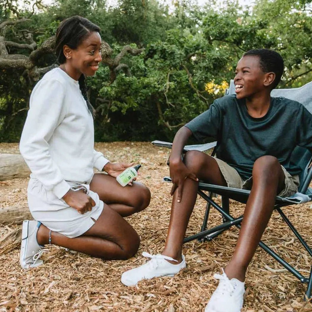 A woman kneels on wood chips holding Natrapel Lemon Eucalyptus Pump 3.4oz while talking to a boy in a chair in a wooded area.
