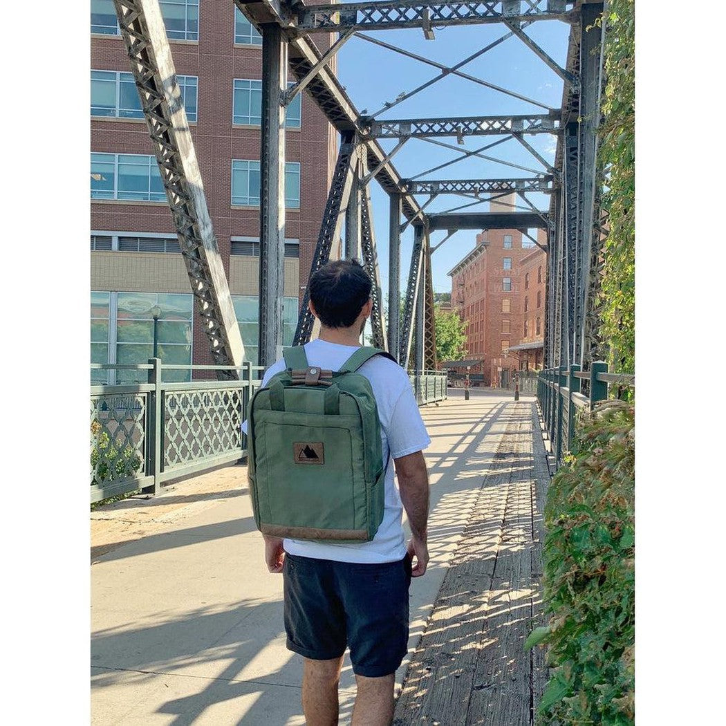 A man in a white tee and black shorts walks on a metal bridge wearing the black Adventurist Backpack Co. Weekender, with brick buildings and greenery in the background.