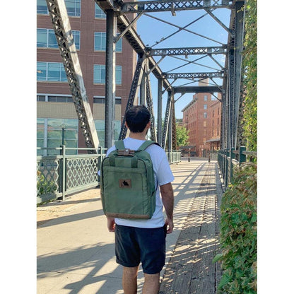 A man in a white tee and black shorts walks on a metal bridge wearing the black Adventurist Backpack Co. Weekender, with brick buildings and greenery in the background.