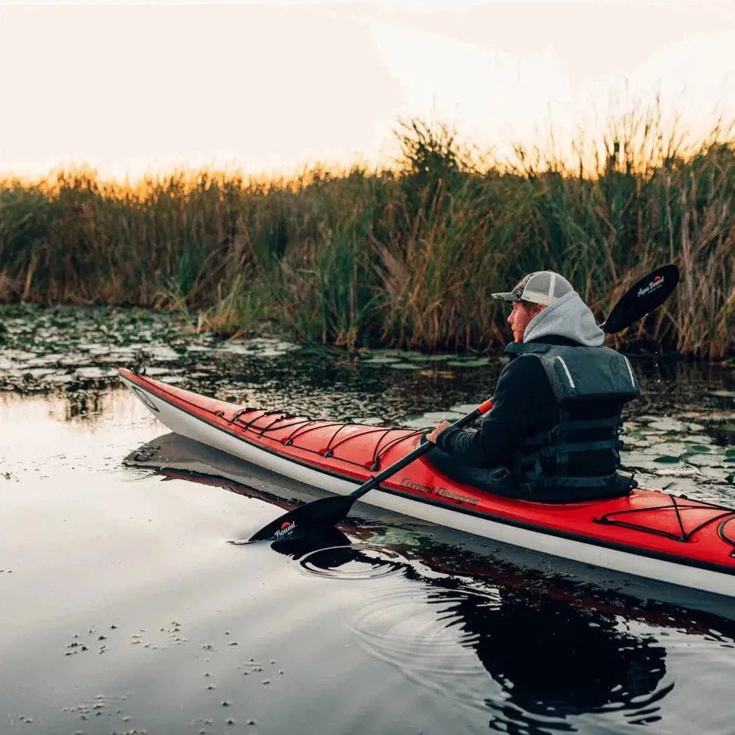 A person in a black life vest sits in a red kayak on calm water with tall grasses in the background, holding a black Aqua Bound Sting Ray Carbon 2-Piece Posi-Lok™ Kayak Paddle.