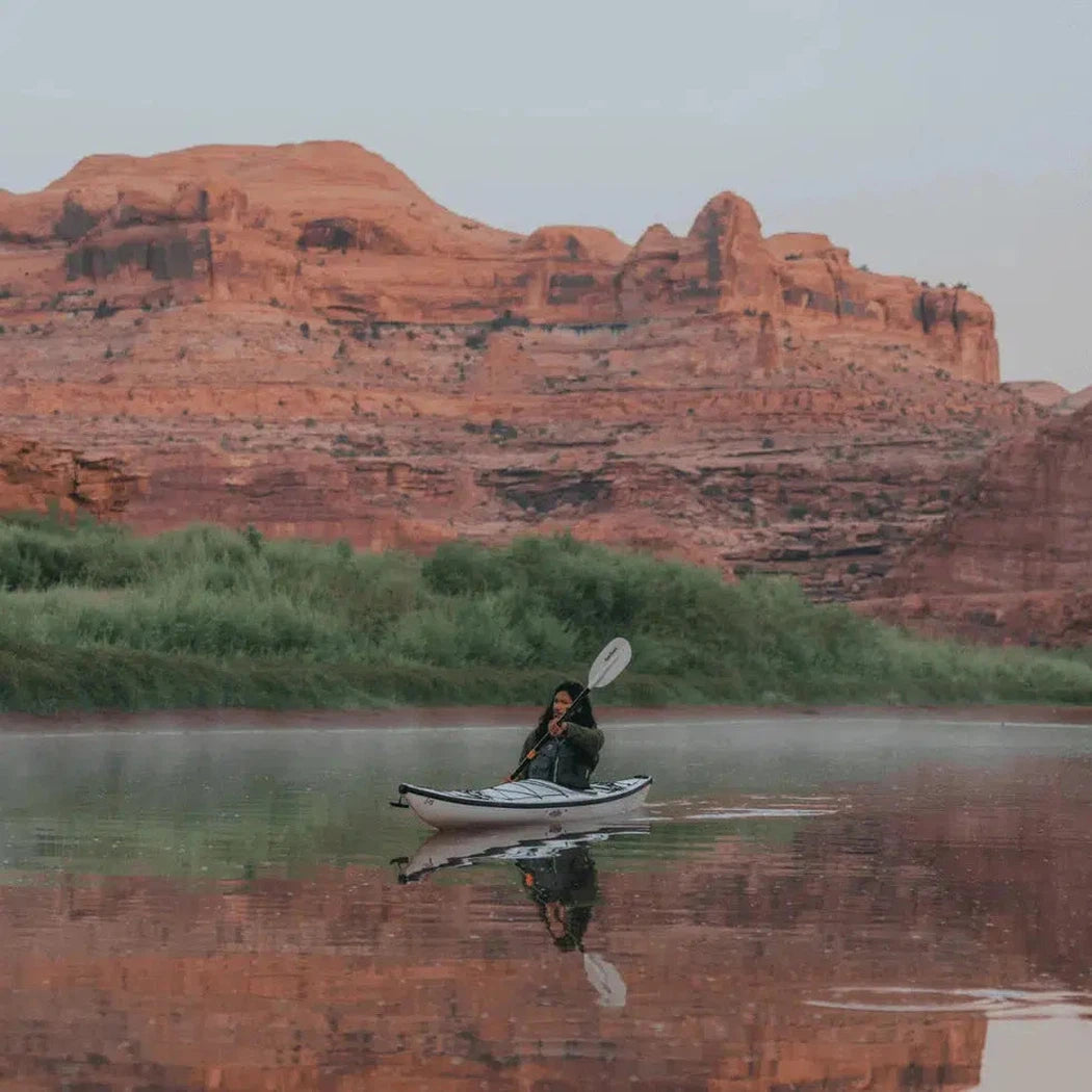 A person kayaks on calm water with red rock formations in the background, using the white Aqua Bound Sting Ray Hybrid 2-Piece Posi-Lok™ Kayak Paddle.
