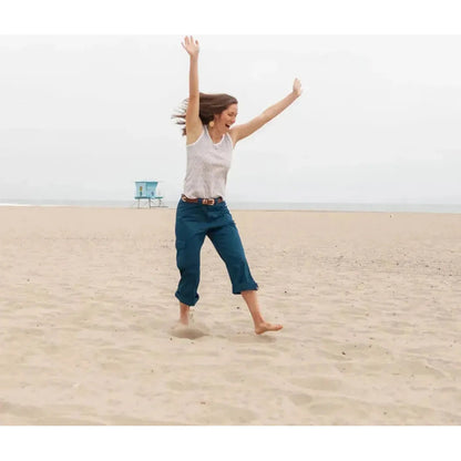 A woman wearing the Aventura Women's Bali Tank in Simply Taupe jumps on a sandy beach with a lifeguard chair in the background.