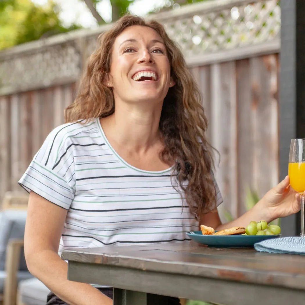 A woman wearing the Aventura Women's Endless Summer Top in Granite Green sits at an outdoor table with a plate of food and a glass of orange juice.