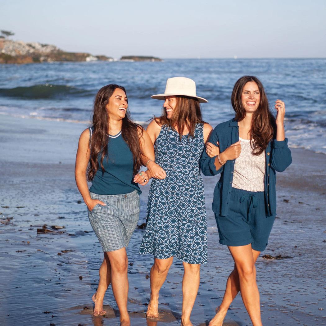 Three women walk barefoot on a beach wearing the Aventura Women's Kelford Full Button in Gibraltar Sea, with the ocean and shoreline in the background.