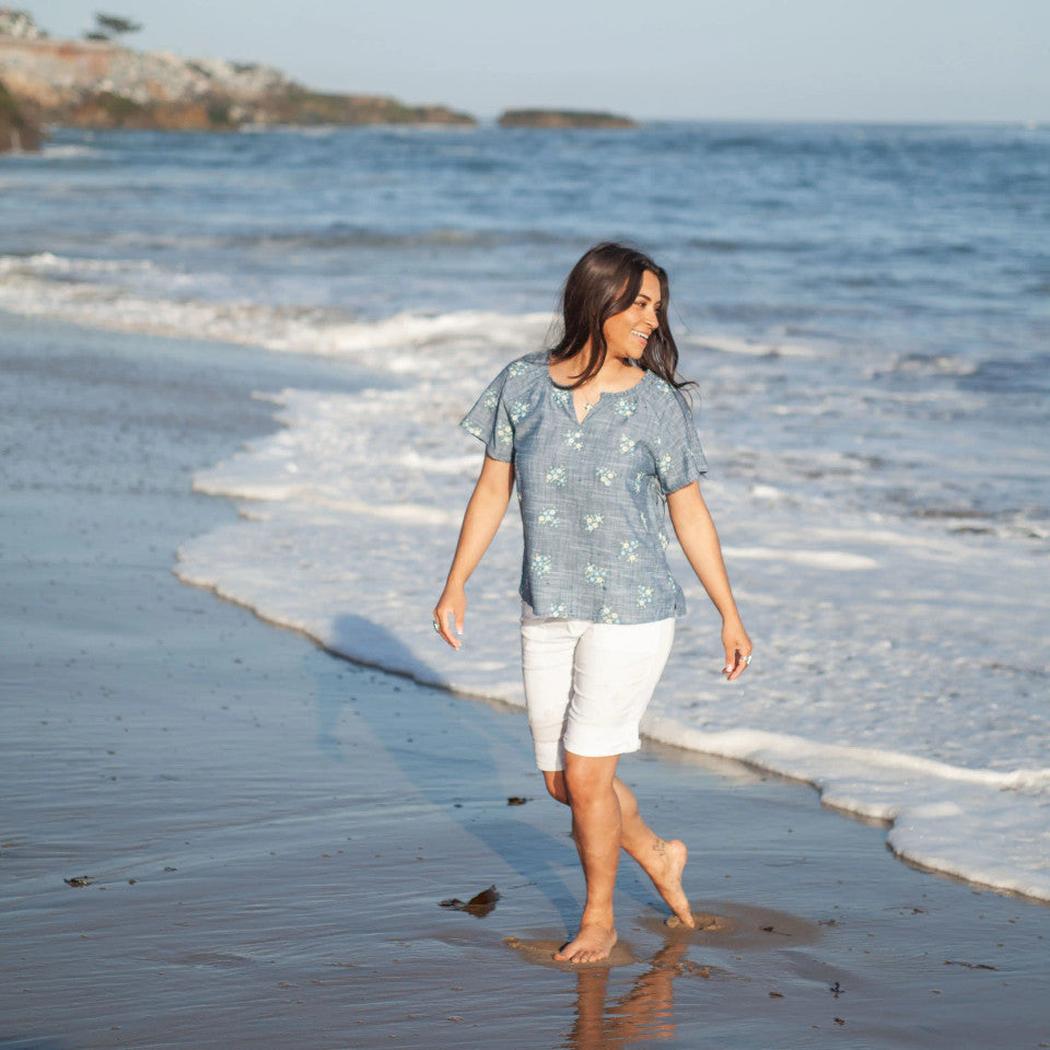 A woman wearing the Aventura Women's Mariposa Top in Gibraltar Sea walks on a beach with ocean waves in the background.
