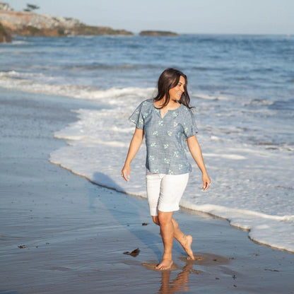 A woman wearing the Aventura Women's Mariposa Top in Gibraltar Sea walks on a beach with ocean waves in the background.