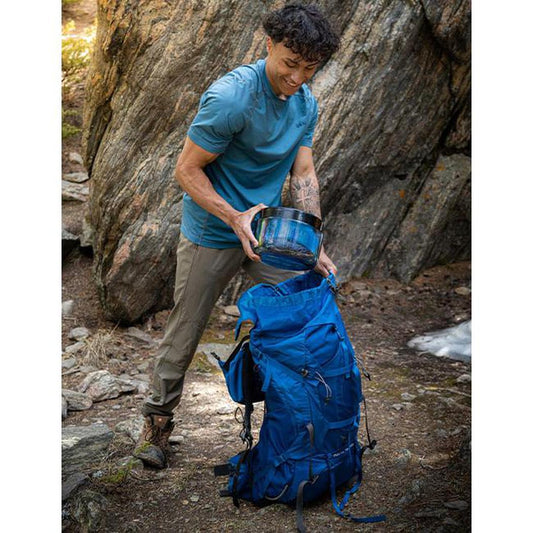 A man puts a Bear Vault BV425 - Sprint into a blue backpack near a large rock.