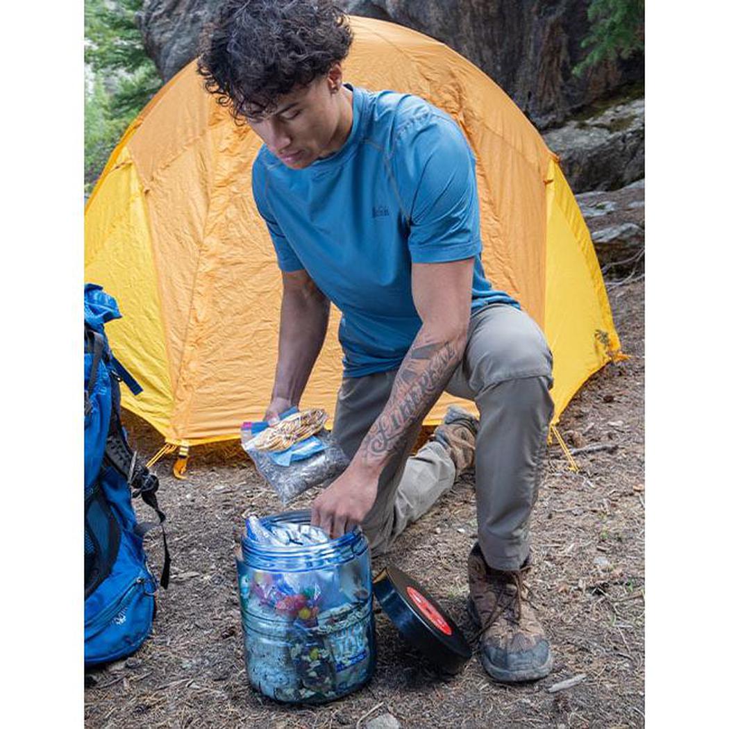 A person kneels by a yellow tent, opening a Bear Vault BV475 - Trek food storage container on the ground.